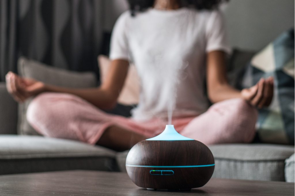 Woman with curly hair sitting on sofa in Lotus pose and meditating with closed eyes near smart air humidifier at home.