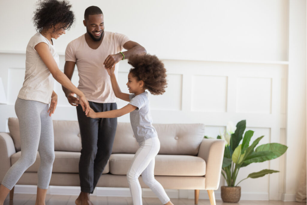 Happy parents dancing with their daughter in the living room, having fun at home while being engaged in physical activity.