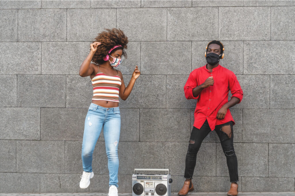 Young African people wearing face mask dancing outdoor while listening to music with wireless headphones and vintage boombox