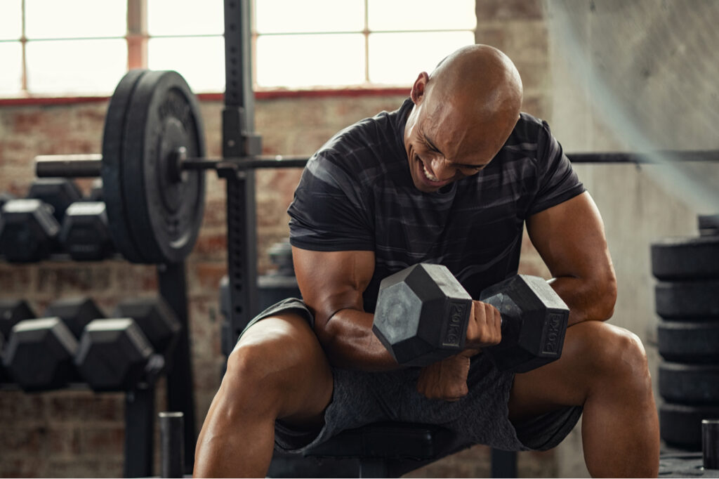Muscular guy lifting dumbbell as a Home gym essentials while sitting on bench at gym.