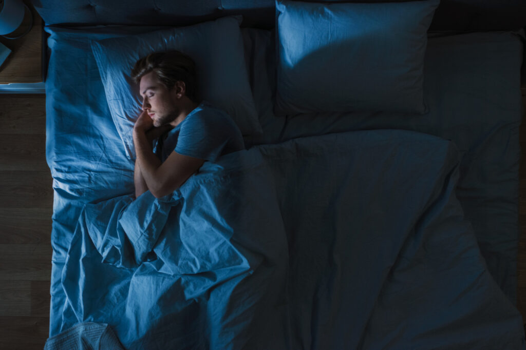 Top View of Handsome Young Man Sleeping Cozily on a Bed in His Bedroom at Night. Blue Nightly Colors with Cold Weak Lamppost Light Shining Through the Window.