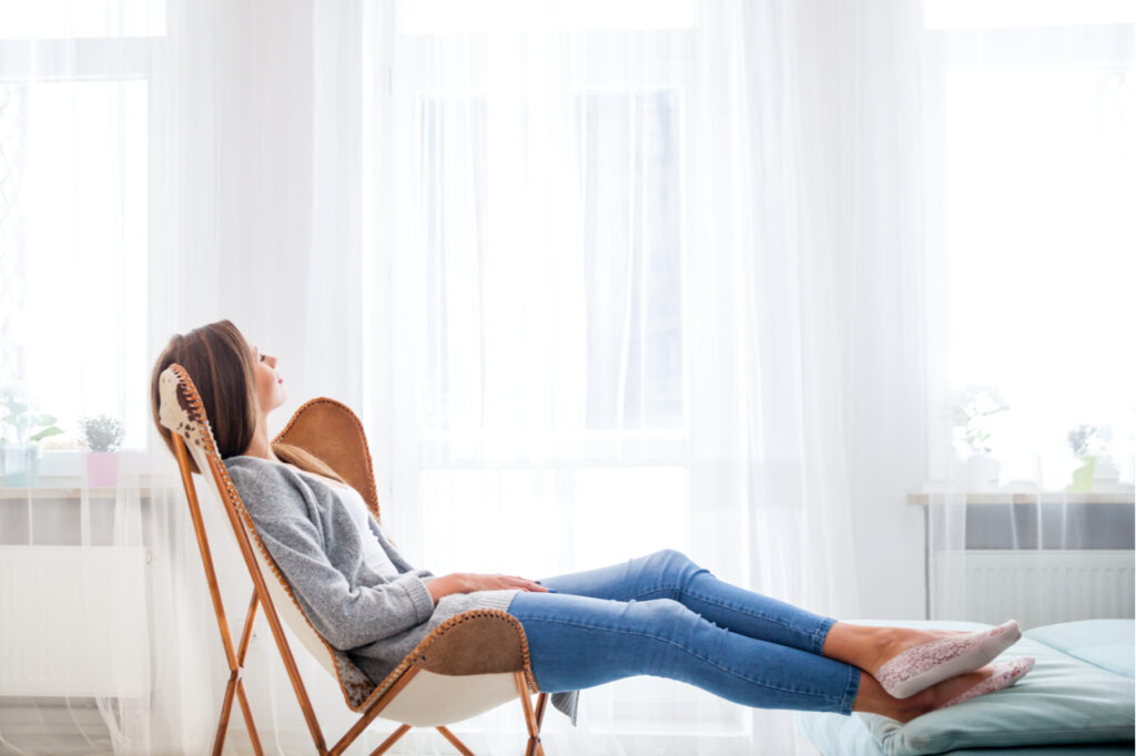 Woman at home sitting on modern chair near window relaxing in living room.