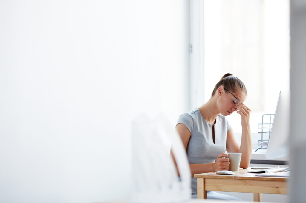 Tired young woman sitting at computer desk with cup of coffee touching forehead with her hand feeling left out.
