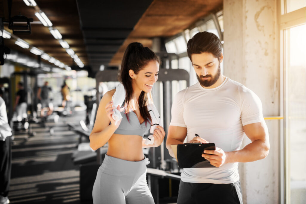 Young slim fitness girl standing with a towel near handsome trainer while showing her schedule for next week in the gym. Maintain a schedule may help improve cognitive function.