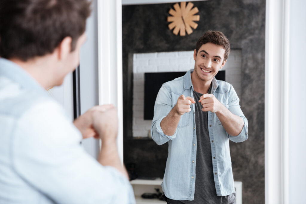 Smiling young man pointing fingers at his mirror reflection while standing at home.