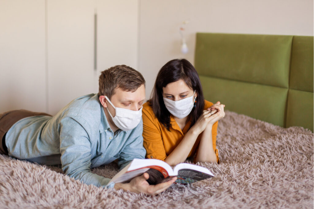 A young couple is sitting at home on self-quarantine. Husband and wife quarantined coronavirus in protective masks. New reality. Normal life in isolation. Read a book together.