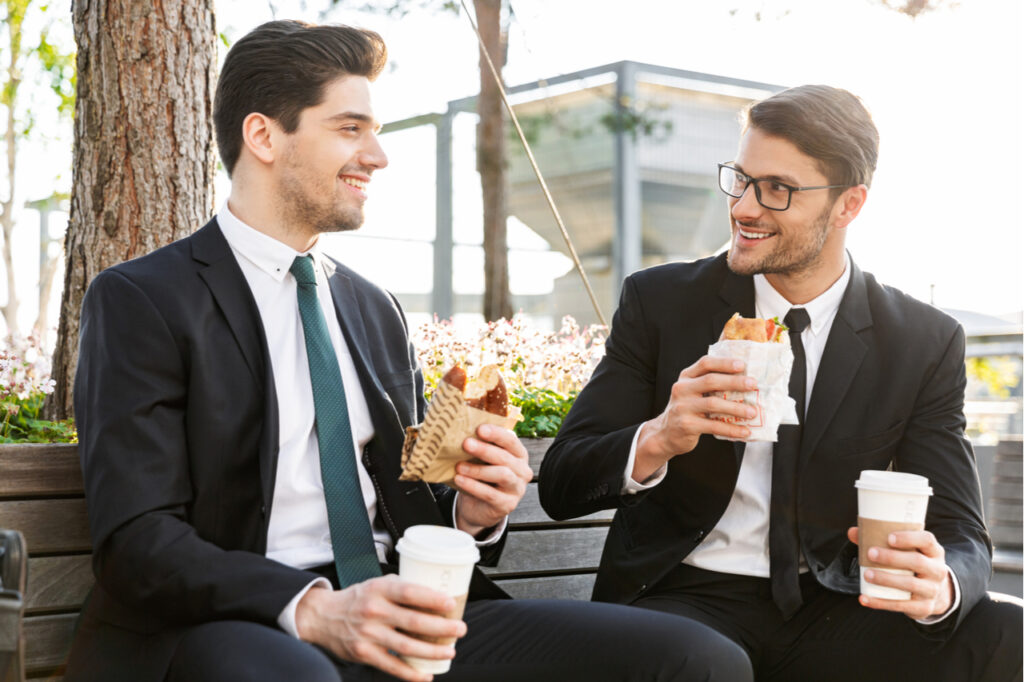 Two attractive smiling young businessmen wearing suits talking while having a lunch break outdoors at the city streets, drinking coffee