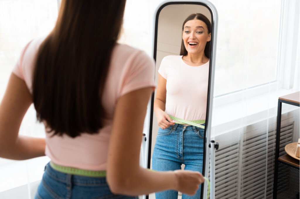 Weight Loss. Happy Girl Measuring Waist With Tape Standing In Front Of Mirror Slimming At Home