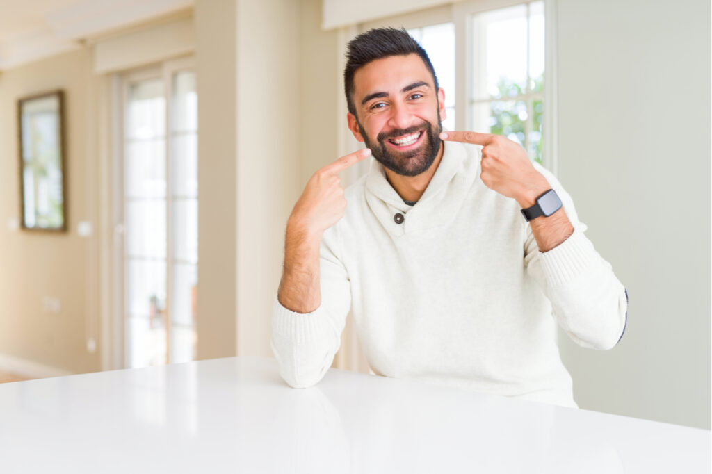 Handsome hispanic man wearing casual white sweater at home smiling confident showing and pointing with fingers teeth and mouth. Health concept.