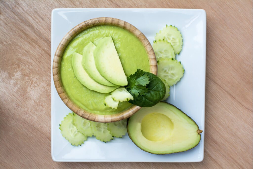 Cucumber avocado soup in bamboo bowl on white square plate on wood background