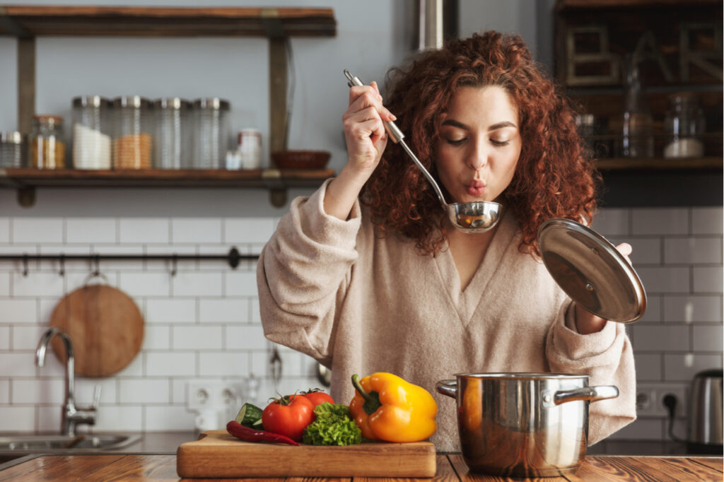 Photo of pretty caucasian woman holding cooking ladle spoon while eating soup with fresh vegetables in kitchen at home. What is your favorite healthy winter recipe?