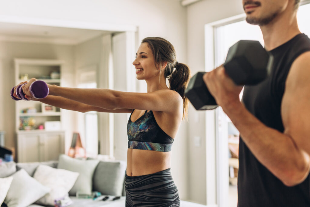 Healthy young couple doing exercises with dumbbells at home. Fit young woman with man doing weights workout indoors in living room.