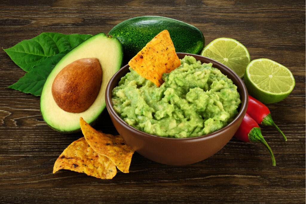 Guacamole in a bowl and nachos on the side, avocado sliced in half, sliced lime and 2 chilis.