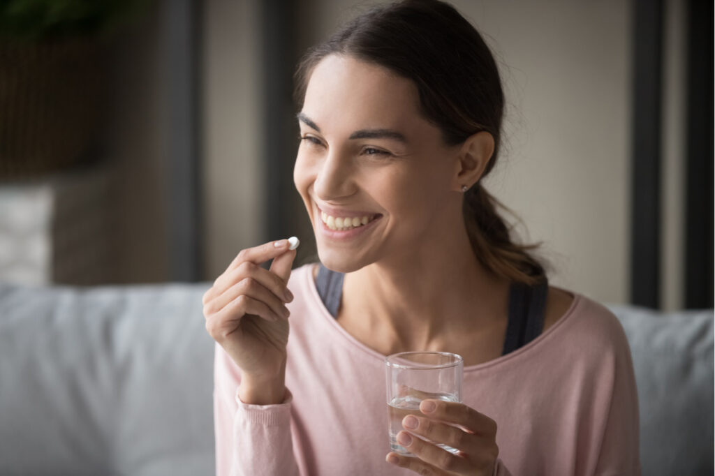Woman holding now foods l-carnitine tartrate and glass of water sit on sofa at home.