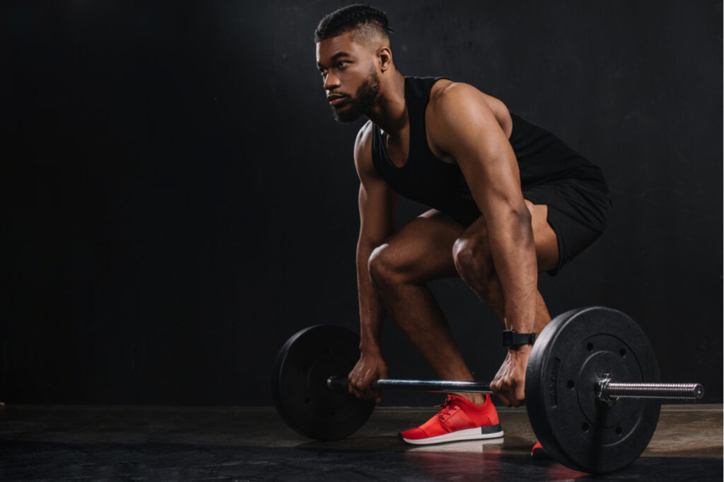 Muscular young sportsman lifting barbell and looking away on black.