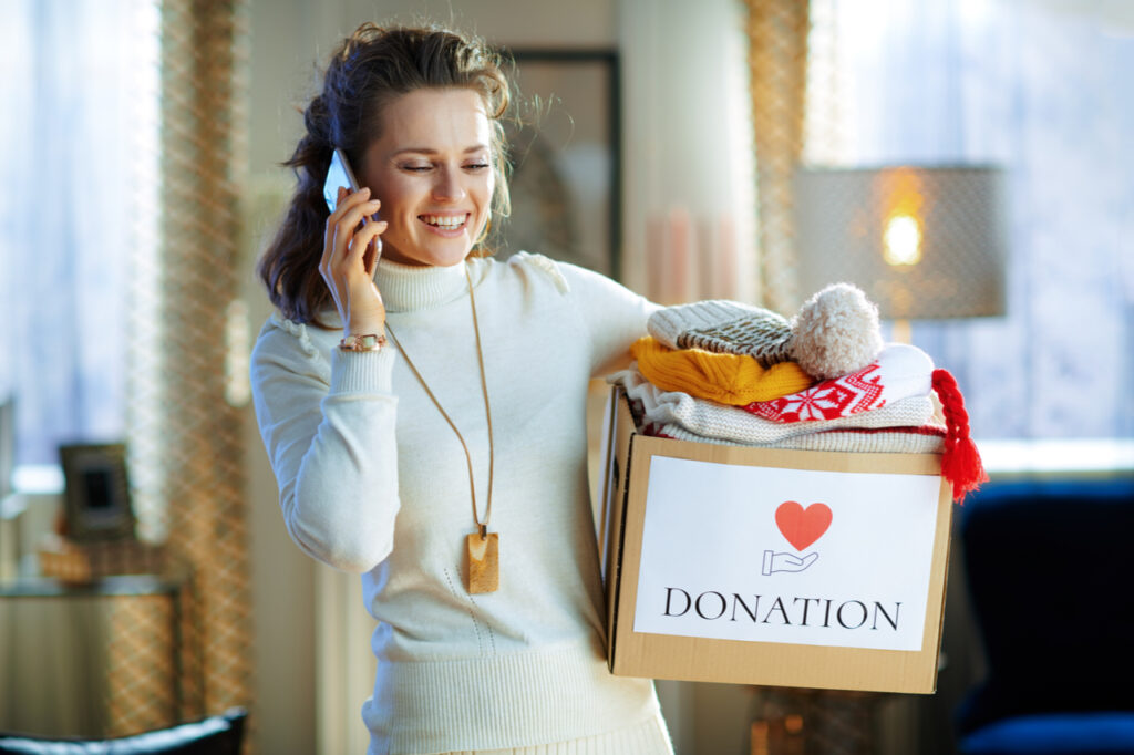 Woman in white sweater and skirt with donation box with old warm clothes talking on a cell phone at modern home in sunny winter day.