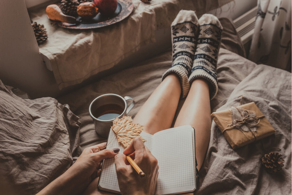 Woman with cup of hot drink sitting by the window and writing in notepad.