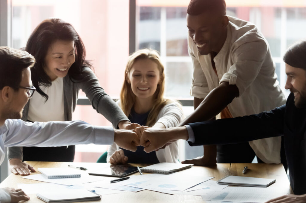 Happy motivated young and older generation multiracial business people joining fists, showing support and team spirit together, celebrating corporate achievement or success at meeting in office.