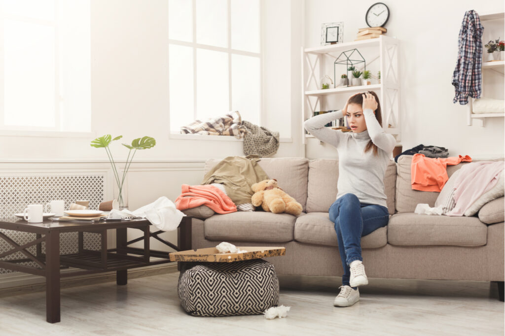 Desperate helpless woman sitting on sofa in messy living room. Young girl surrounded by many stack of clothes. Her physical environment is messy.