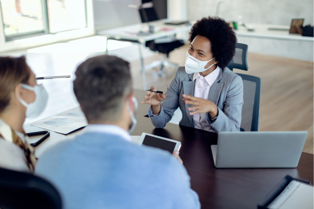 Happy black financial advisor communicating with a couple on a meeting in the office. All of them are wearing protective face masks due to COVID-19 pandemic.