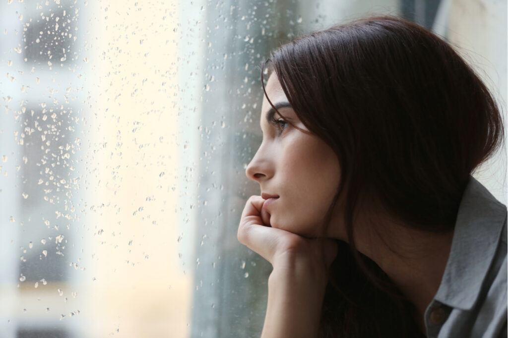 Depressed young woman near window at home with seasonal affective disorder.