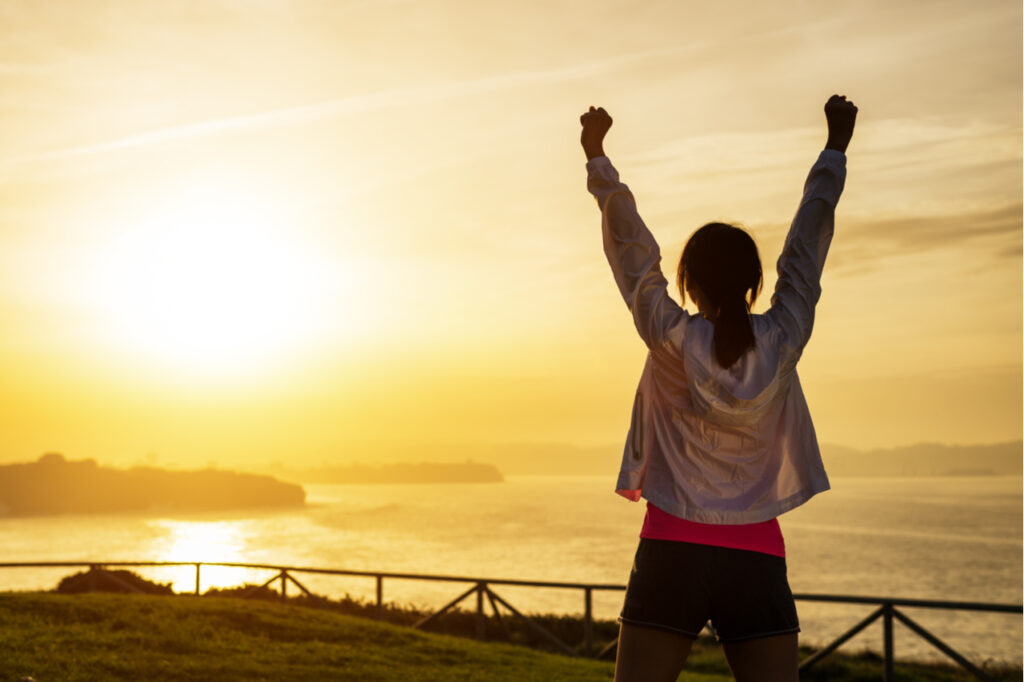 Successful sporty woman raising arms towards golden beautiful sunset as a way on how to motivate yourself to work out.