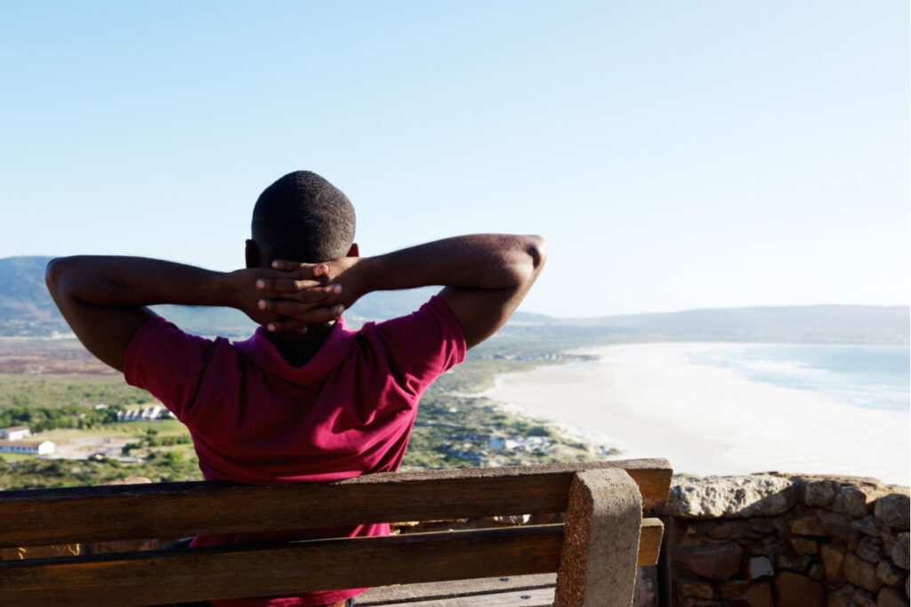 Rear view portrait of young african man sitting relaxed on a bench with his hands behind head, young guy on vacation.