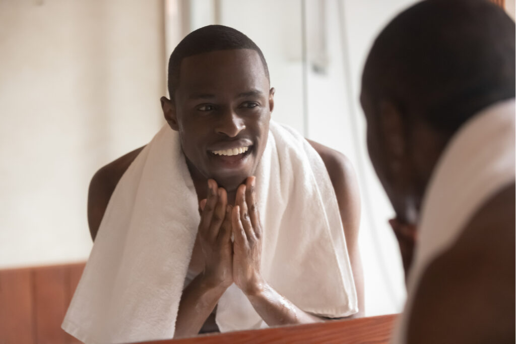 Man with a towel on shoulders standing front of the mirror knows how to get rid of acne fast.