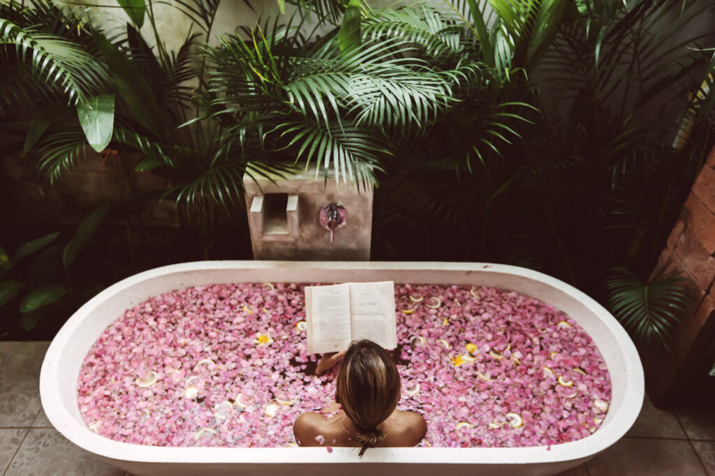 Woman reading book while relaxing in bath tub with flower petals.