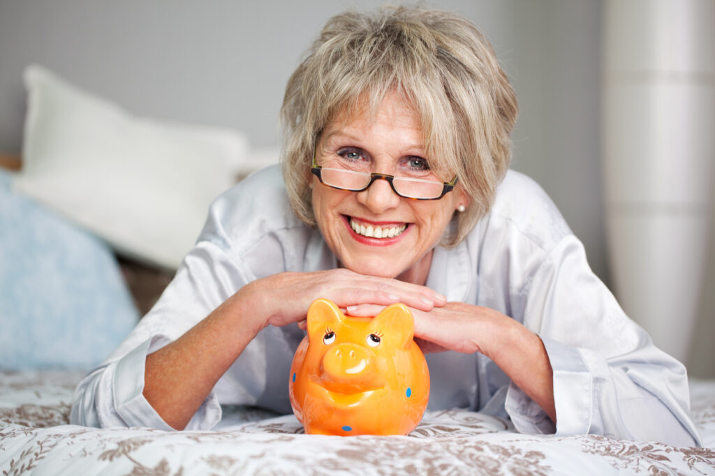 Conceptual portrait of happy senior female lying on bed with piggybank.