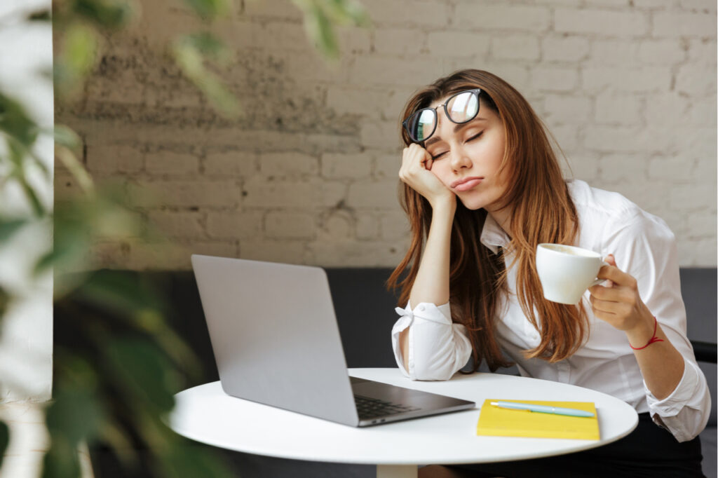 Portrait of a tired young businesswoman sitting at the table with laptop computer while holding cup of coffee.