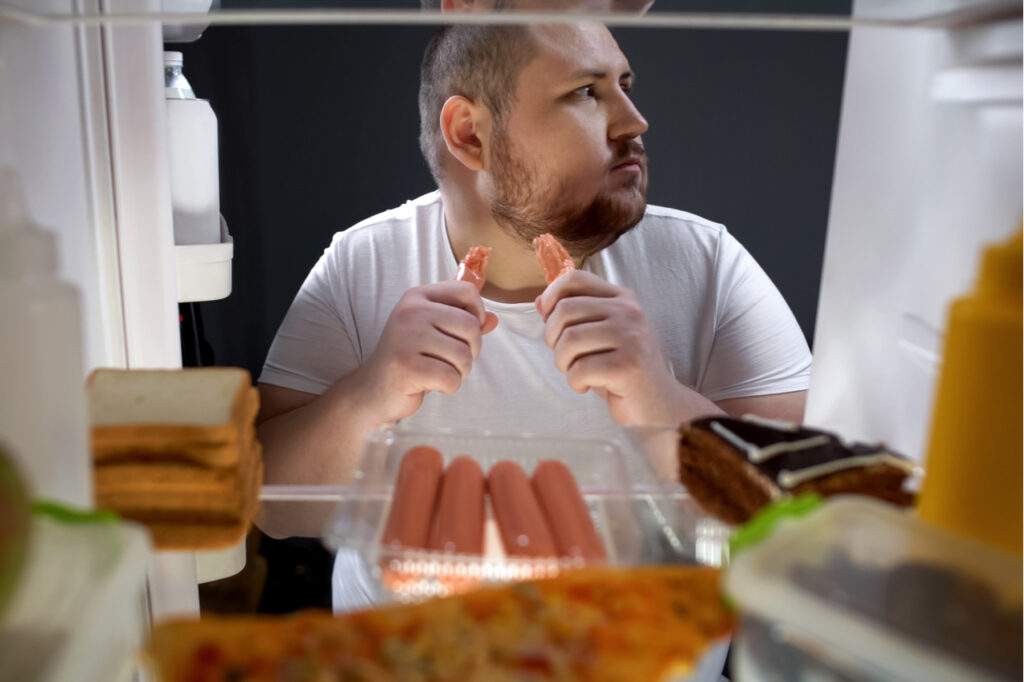 Addicted man secretly eating sausages at night near fridge, unhealthy nutrition.