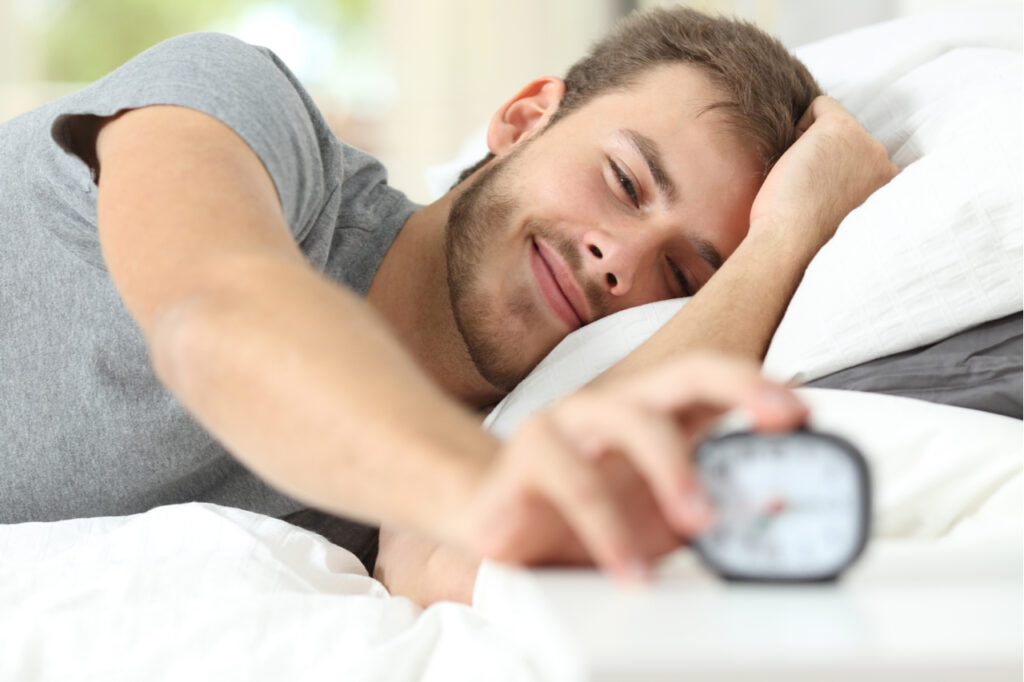 Happy wake up of a happy man lying on the bed and stopping alarm clock.