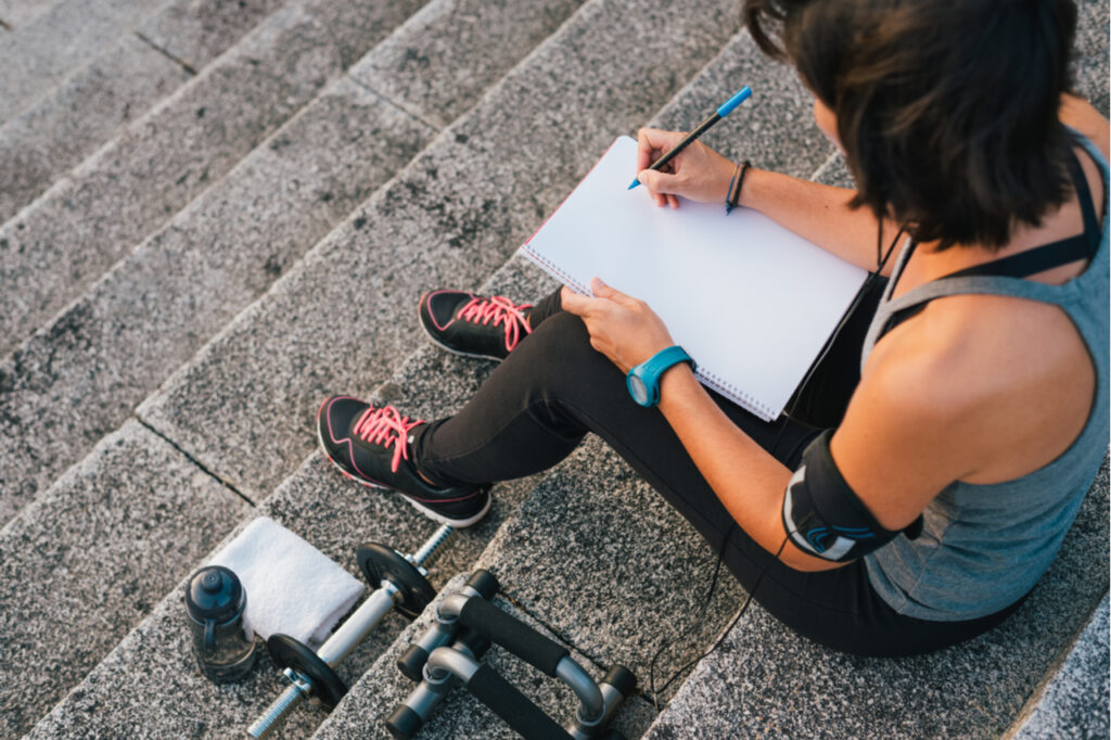 Fitness sporty woman writing on blank notepad while sitting on urban stone stairs before exercises workout routine.