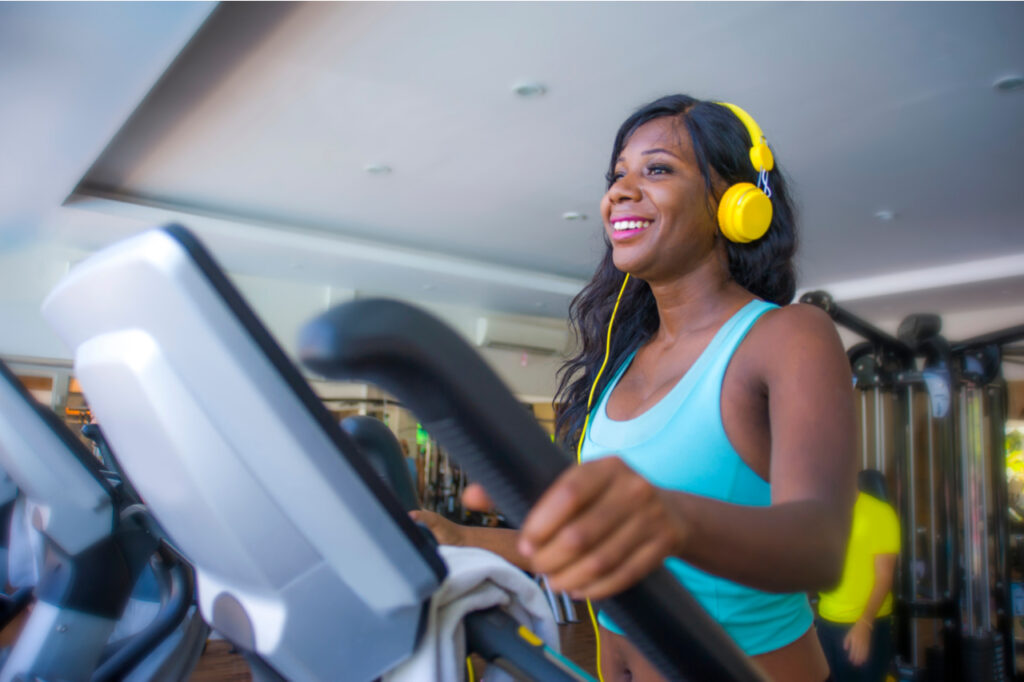 indoors gym portrait of young attractive and happy black african American woman with headphones training elliptical machine workout at fitness club smiling cheerful in healthy lifestyle
