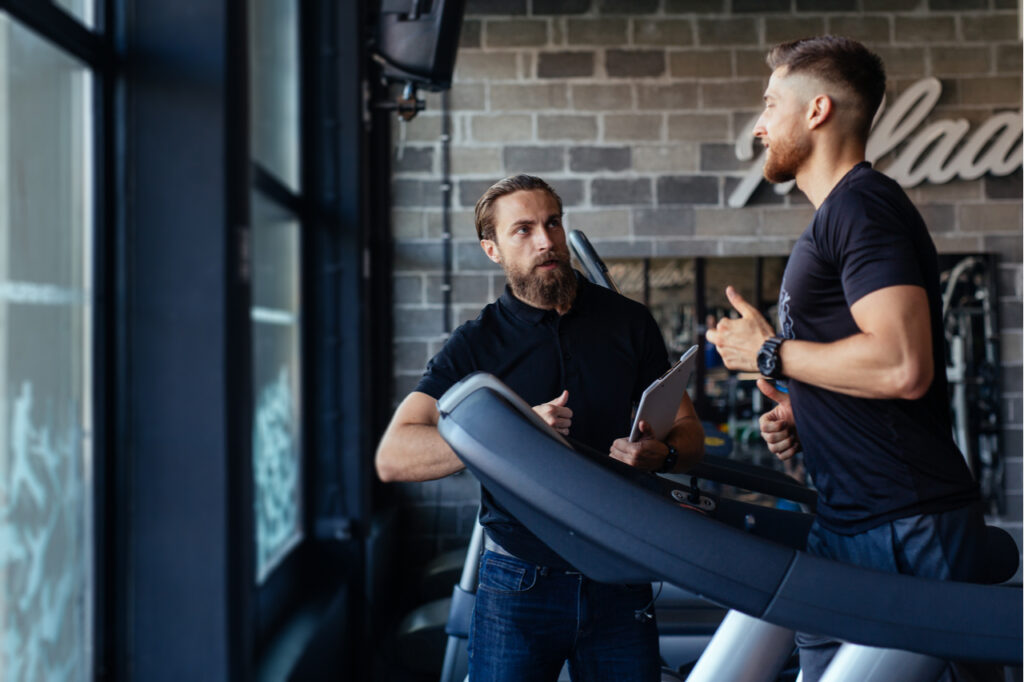 A personal trainer and a man on a treadmill in the gym talking about how to become a certified personal trainer.