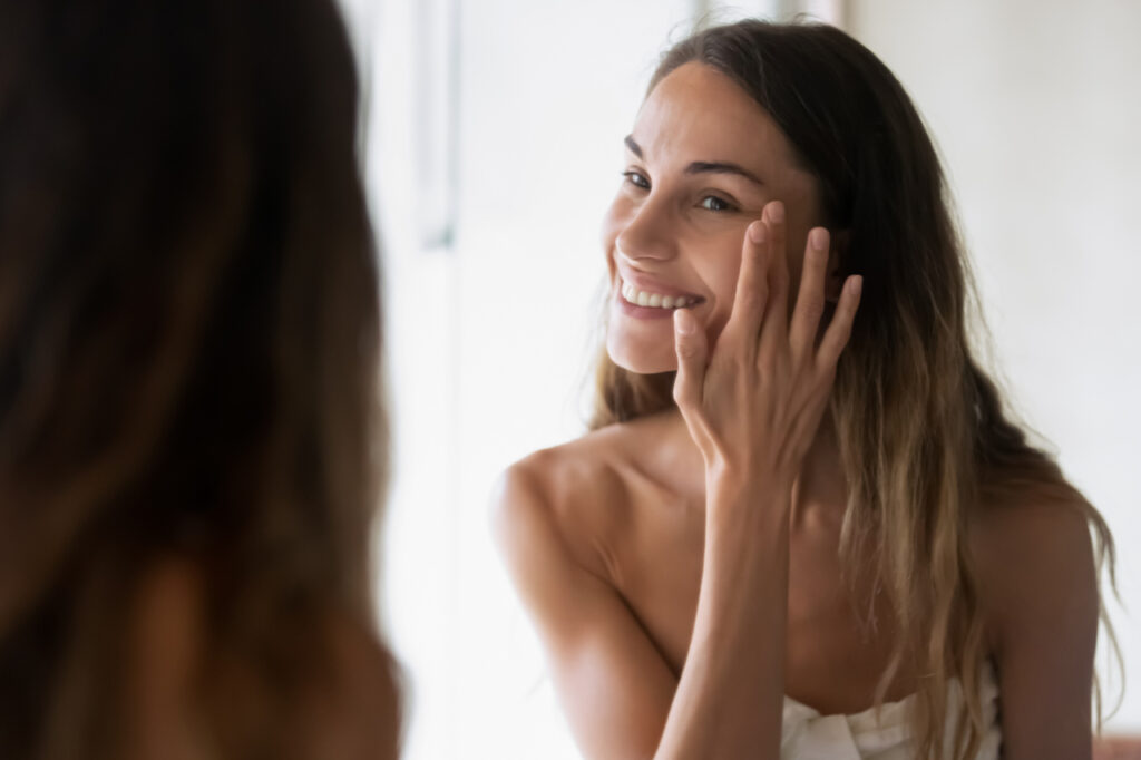 Smiling girl in bath towel look in mirror apply moisturizing facial cream or serum, happy young woman perform daily morning skincare face routine for healthy glowing skin, do beauty procedure