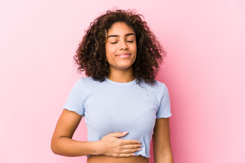 Young african american woman against a pink background touches tummy, indicating good gut health.