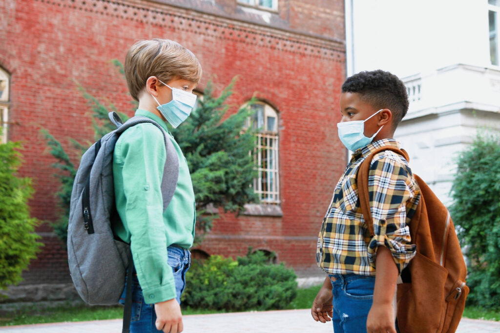 Portrait of mixed-races little boys in masks greeting each other in front of school in quarantine thinking of outdoor activities for kids.