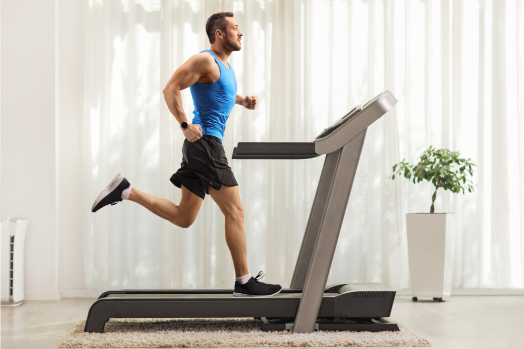 Full length profile shot of a young man running on a treadmill at home