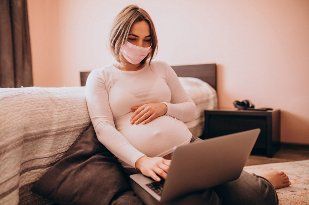 Pregnant woman wearing mask having a video chat with family on a laptop.