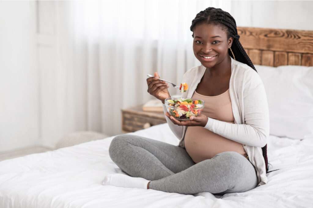 Happy pregnant woman eating fresh vegetable salad on bet at home.