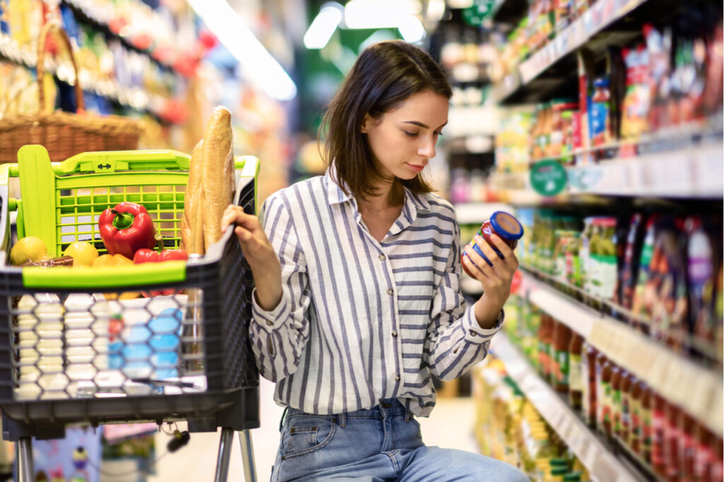 Young Woman With Shopping Cart In Market Buying Groceries Food Taking Products From Shelves In Store, Holding Glass Jar Of Sauce, Checking Label Or Expiry Date