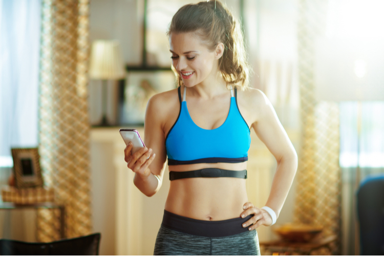 Smiling young sports woman in fitness clothes in the modern living room wearing heart rate monitor using smartphone to track heart rate in fitness app.