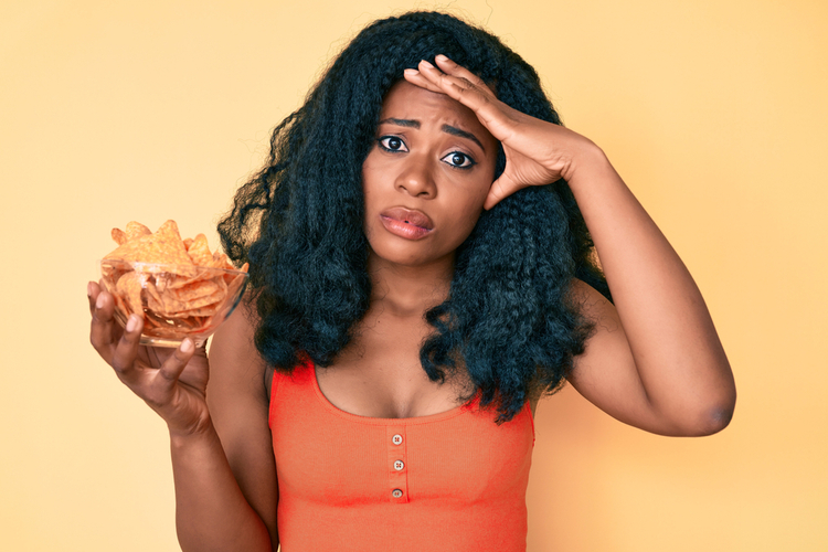 Woman holding nachos potato chips stressed and frustrated with hand on head.