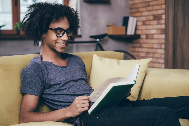 Man reading positive affirmations journal with smile and wearing glasses while sitting on the couch at home.