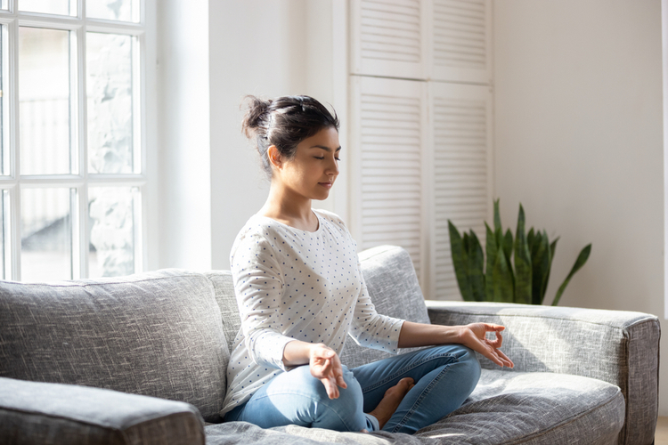 Female sitting on couch lotus pose put hands on lap folded fingers closed eyes.