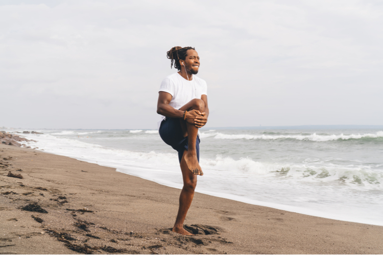 Male athlete in sportswear standing on sandy beach and doing stretching exercise during workout in summer.
