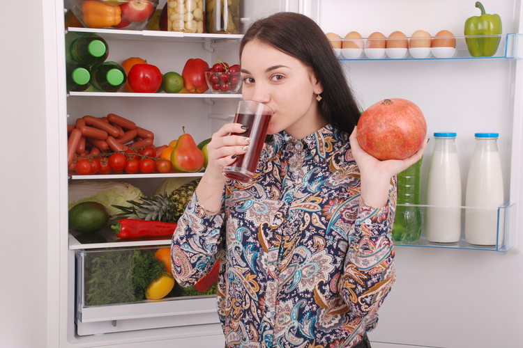 Woman holds a glass of pomegranate juice and garnet on the fridge background.