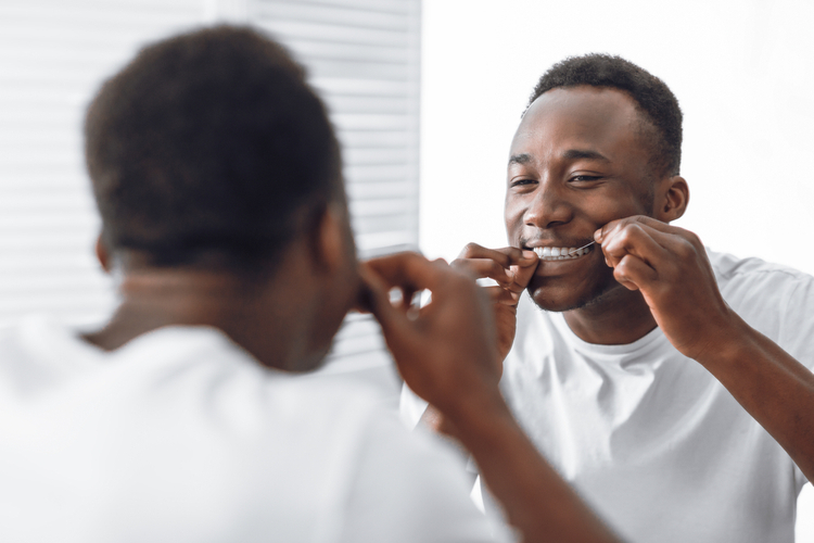 Young man flossing his teeth in the bathroom while looking at the mirror.